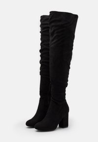 Botas de ante negras por encima de la rodilla con tacones bloque y cremalleras laterales, exhibidas de pie sobre un fondo blanco.