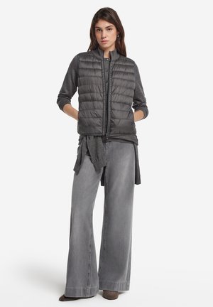 Gilet en duvet gris avec des coutures horizontales, fermeture éclair et col haut, porté par-dessus un haut gris et associé à un jean gris à jambes larges.