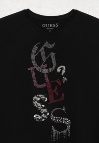 Schwarzes T-Shirt mit mehrfarbigen Pailletten- und Strassakzenten, die das Wort "GUESS" in verschiedenen Größen und Stilen auf der Vorderseite darstellen.