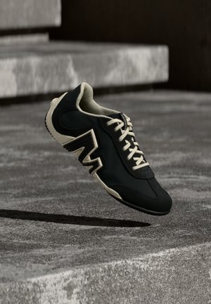 Basket sneaker noire et beige avec semelle texturée et grand détail "M", flottant au-dessus d'escaliers en béton texturé dans un éclairage neutre.