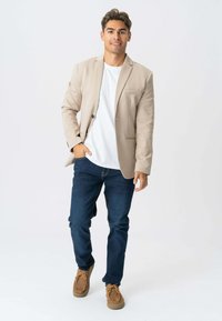 Beige blazer med to knapper og frontlommer, båret over en hvid T-shirt, parret med mørkeblue jeans og brune sko.