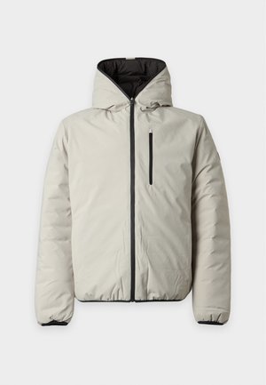 Jeune homme debout portant une veste beige à capuche sur une chemise noire, un pantalon noir et des chaussures marron foncé sur un fond blanc uni.