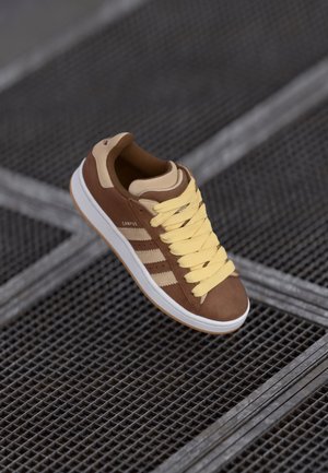 Bruine en beige sneaker met gele veters en witte zool, gelabeld "Campus", geplaatst op een metalen roostervloer.