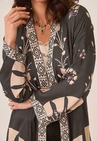 Mujer vestida con un kimono de estampado floral en negro y beige con ribete geométrico y un collar de cuentas doradas con piedras moradas y azules.