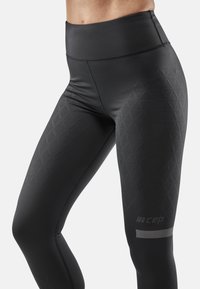 Leggings noirs taille haute avec une texture lisse, présentant un motif en diamant sur le côté et un accent de logo sur le bas de la jambe.