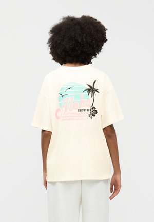 Άτομο με σγουρά μαλλιά που φοράει μια κρεμ μπλούζα με παστέλ σχέδιο "Aloha Surf Club", φοινικόδεντρο και πουλιά στην πλάτη.