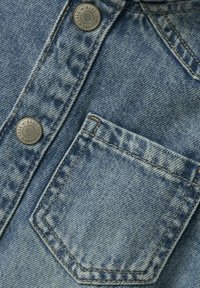 Svijetloplava traper tkanina s izblijedjelom teksturom. Ima metalne kopče i zakrpa džep na lijevoj strani, s vidljivim detaljima šivanja.