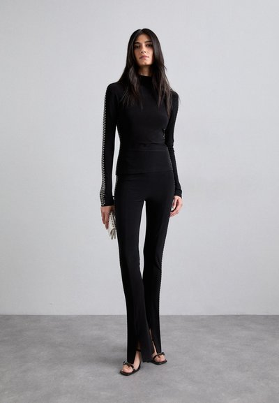 Black long-sleeve top with open knit detailing along the sleeves, paired with fitted black pants featuring side slits. Textured fabric throughout.