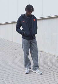 Sudadera negra con cremallera y logo naranja, camiseta negra, pantalones gray texturizados anchos y zapatillas multicolor sobre un fondo de pavimento de baldosas.