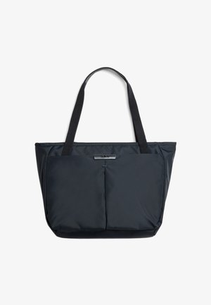 Borsa tote nera con due tasche frontali piatte, spallacci e una piccola targhetta rettangolare con logo sopra le tasche su sfondo bianco.