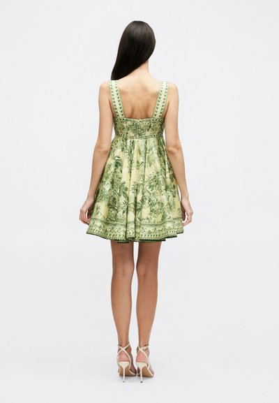 Γυναίκα με μακριά σκούρα μαλλιά, που φοράει κοντό πράσινο φόρεμα με λουλουδάτο μοτίβο χωρίς μανίκια και μπεζ ψηλοτάκουνα σανδάλια, στέκεται γυρισμένη με την πλάτη.