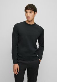 Schwarzer Strickpullover mit langen Ärmeln und Rundhalsausschnitt. Mit einem dezenten Logo auf der Brust und einer taillierten Taille. Glatte Textur.