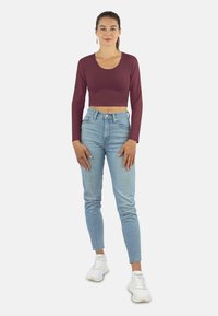 Burgunderfarbenes langärmliges Crop-Top kombiniert mit hellblauen hochgeschnittenen Jeans und weißen Sneakers, stehend mit Händen auf den Hüften vor einem schlichten Hintergrund.