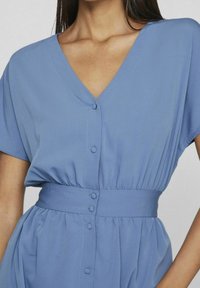 Vaaleansininen mekko, jossa on lyhyet hihat, v-pääntie ja tiukka vyötärö. Edessä nappikirjailua ja kerättyä tekstuuria vyötäröllä.