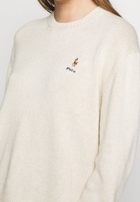 Cremefarbener, gestrickter Pullover mit Rundhalsausschnitt. Verfügt über ein kleines gesticktes Logo eines Reiters und das Wort "Polo" in Blau.