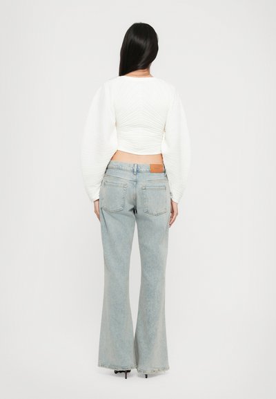 Witte cropped top van gestructureerde stof; lichtblauwe flare jeans met achterzakken, een bruine leren label op de tailleband en een vervaagde afwerking.