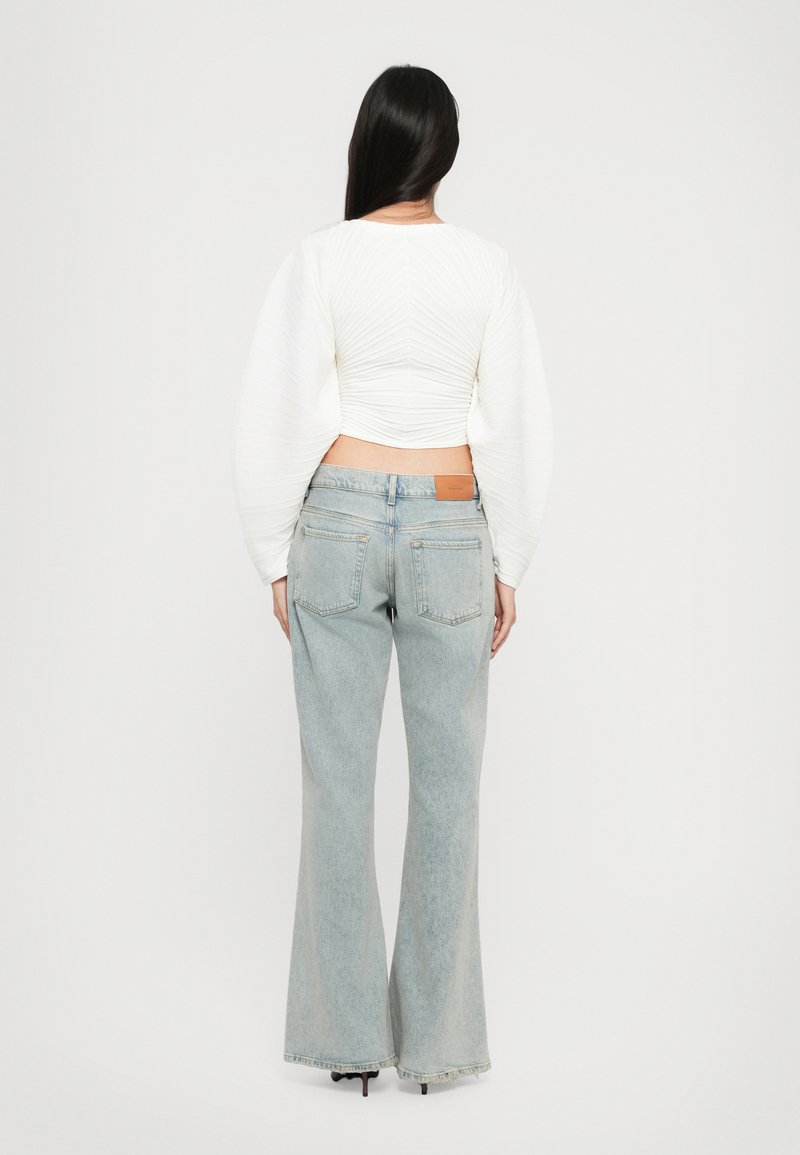 Top court blanc en tissu texturé ; jean évasé bleu clair avec poches arrière, étiquette en cuir marron sur la taille et finition délavée.
