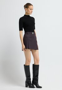 Kvinde iført en tætsiddende sort turtleneck med korte ærmer, lilla stribet miniskørt med bælte og sorte knæhøje støvler med hæl, står med siden til.