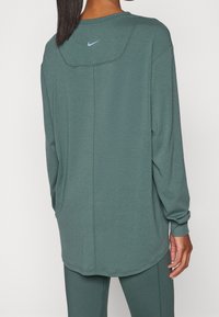 Grön långärmad topp med rundad halsringning, med en sydd detalj på ryggen och en liten blå Nike-logotyp nära kragen.