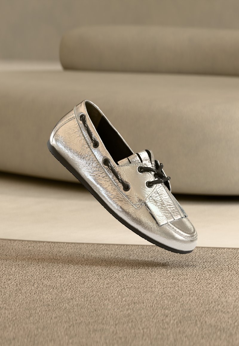 Μοκασίνι σε μεταλλικό ασημί χρώμα με υφή στην επιφάνεια, μαύρα κορδόνια και μαύρες λεπτομέρειες. Στρογγυλεμένη μύτη και επίπεδη λαστιχένια σόλα.