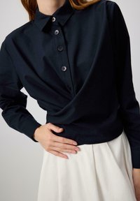 Camicia blu navy con bottoni e maniche lunghe, caratterizzata da un collo a camicia e un dettaglio frontale attorcigliato. Tessuto morbido abbinato a una gonna bianca.