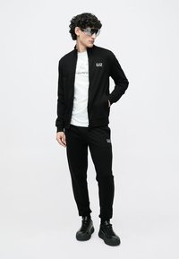 Schwarzer sportlicher Trainingsanzug mit einer Reißverschlussjacke mit Taschen, kombiniert mit passenden Hosen. Das Material hat eine glatte Textur; Logo-Akzente sind sichtbar.