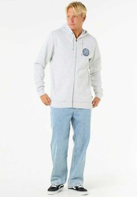 Sweat à capuche zippé gris clair avec une patch logo sur le devant, manches longues, capuche spacieuse et poignets côtelés, associé à un jean en denim bleu clair et des baskets noires.