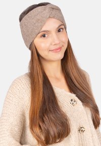 Bandeau en tricot marron avec un design à l'avant torsadé. Porté par un mannequin aux cheveux longs et droits, associé à un gros pull beige.