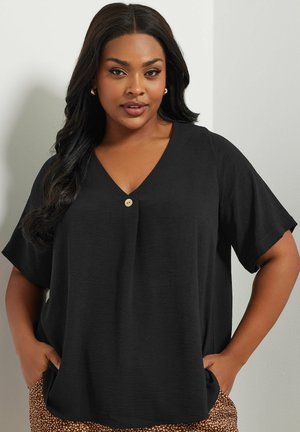 Femme aux longs cheveux foncés portant un chemisier noir à col en V avec un seul bouton et un pantalon marron à motifs, debout les mains dans les poches.