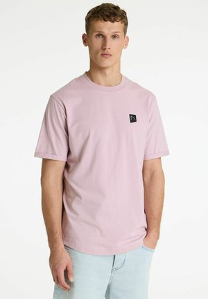 Młody mężczyzna z kręconymi włosami, ubrany w jasnoróżowy t-shirt z krótkim rękawem i jasnoniebieskie jeansy, stojący z jedną ręką w kieszeni.