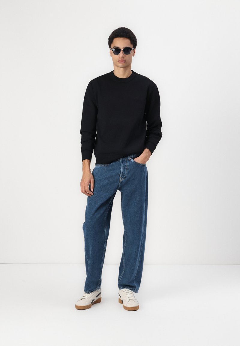 Felpa nera, jeans blu a vestibilità ampia e sneakers bianche con suole marroni. Modello che indossa occhiali da sole scuri, in piedi su uno sfondo semplice.