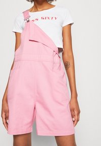 Růžové denimmové lacláče s nastavovacími popruhy, které mají široký a krátký střih a dvě přední kapsy. Stylizováno s bílým zkráceným tričkem.