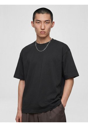 Ung mand med kort hår iført en sort oversize T-shirt, sølvkæde og brune bukser, med den ene hånd i lommen.