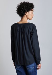 Blusa negra de manga larga con escote redondo, con un detalle de espalda plisada. Combinada con unos vaqueros de mezclilla azul. Textura suave.