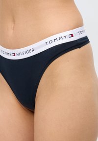 Γυναίκα που φοράει μπλε εσώρουχο στυλ μπικίνι με λευκή μέση που φέρει το λογότυπο Tommy Hilfiger και το έμβλημα της σημαίας.