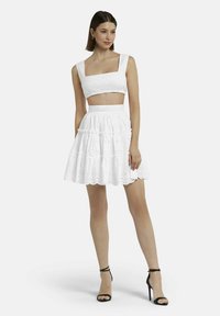 Witte crop top met vierkante halslijn en geborduurde details, gecombineerd met een gelaagde, ruches rok in het wit. Model draagt zwarte sandalen met bandjes.
