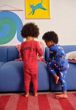 Deux enfants en pyjama Spider-Man sont debout et grimpent sur un canapé bleu dans une pièce colorée avec de l'art mural moderne.