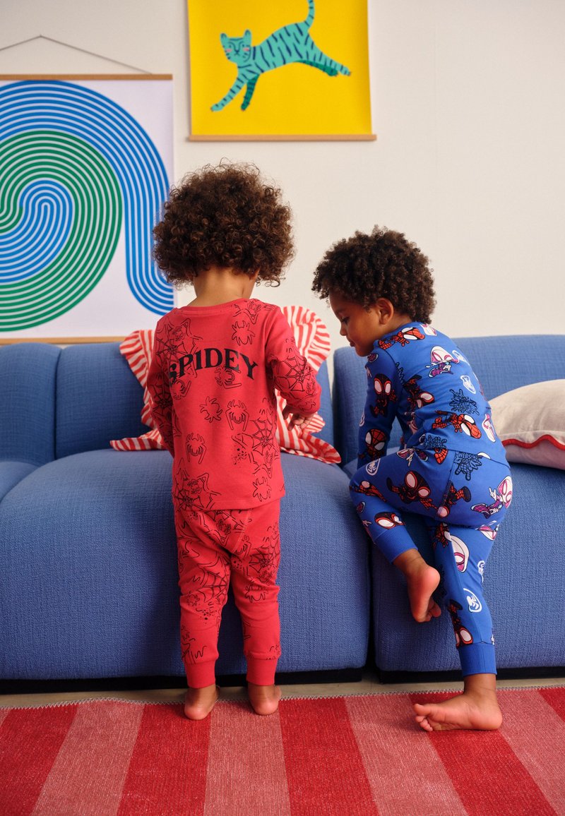 Deux enfants en pyjama Spider-Man sont debout et grimpent sur un canapé bleu dans une pièce colorée avec de l'art mural moderne.