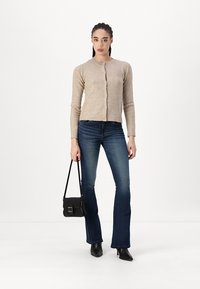 Beige strikkegenser med rund hals og knappelukking, kombinert med mørkeblå utsvungne jeans og en svart crossover-veske.