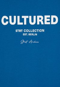 Modrá bavlněná tričko s výrazným bílým textem: "CULTURED" nahoře, s "STRRT COLLECTION EST. BERLIN" a "Street Archive" níže v menším písmu.