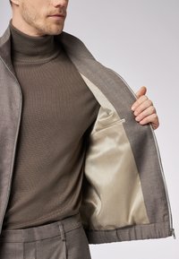 Graue Zip-up-Jacke mit glattem, hellfarbenem Satin-Innenfutter und einer Innentasche. Getragen über einem dunkelbraunen Rollkragenpullover.