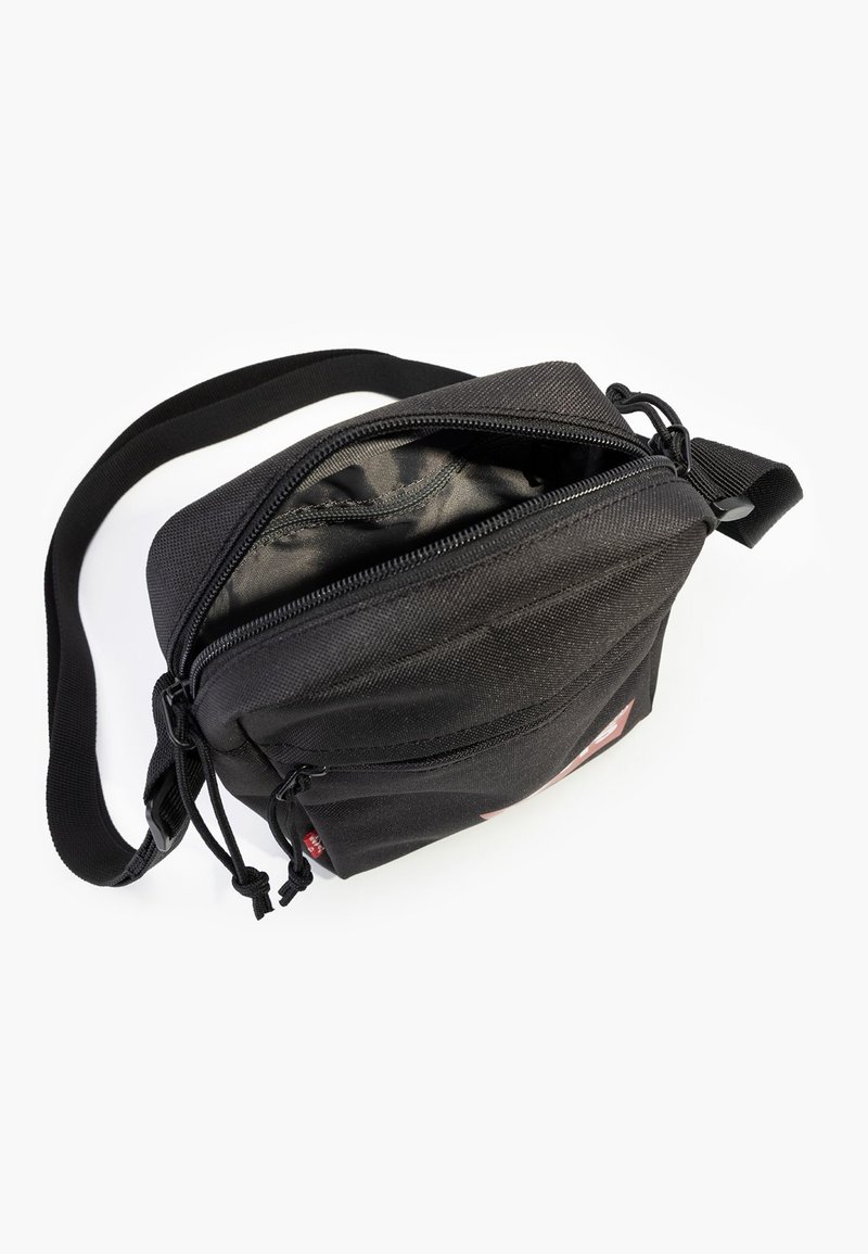 Bolso bandolera de tela negra con cierre de cremallera, que cuenta con una correa ajustable y un pequeño parche con el logo en la parte frontal. Forro interior suave.