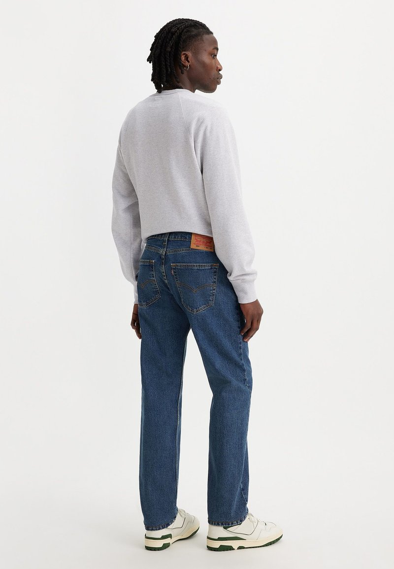 Sweatshirt gris court, jeans droits bleu avec une étiquette rouge sur la poche arrière, et baskets blanches avec des accents verts.