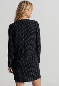 Schwarzes Kleid mit langen Ärmeln, hohem Halsauschnitt und gepunktetem Muster. Der Rücken hat einen verdeckten Reißverschluss und eine lockere Passform.
