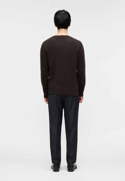 Σκούρο καφέ πλεκτό πουλόβερ με ριμπ τελείωμα, συνδυασμένο με γκρι παντελόνι με λεπτές ρίγες. Το σύνολο χαρακτηρίζεται από άνετη γραμμή και ομαλή υφή.