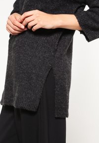 Pull en maille gris foncé avec une coupe décontractée, manches retroussées, fentes latérales, tissu texturé, porté avec un pantalon noir.