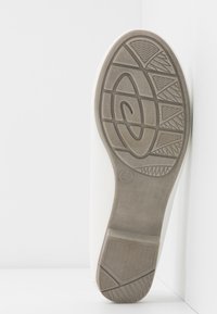 Semelle d'une chaussure blanche avec une bande de roulement en caoutchouc gris présentant des motifs géométriques et une surface texturée antidérapante, inclinée contre un fond blanc d'angle.