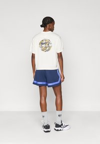 Κοντομάνικη κρέμα μπλούζα με πολύχρωμο γραφικό στο πίσω μέρος, σε συνδυασμό με ναυτική μπλε σορτς που διαθέτουν μια ανοιχτού μπλε λωρίδα.