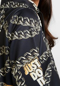 Black hoodie featuring a gold chain pattern and a white "JUST DO IT" graphic. Material appears smooth with a relaxed fit design.