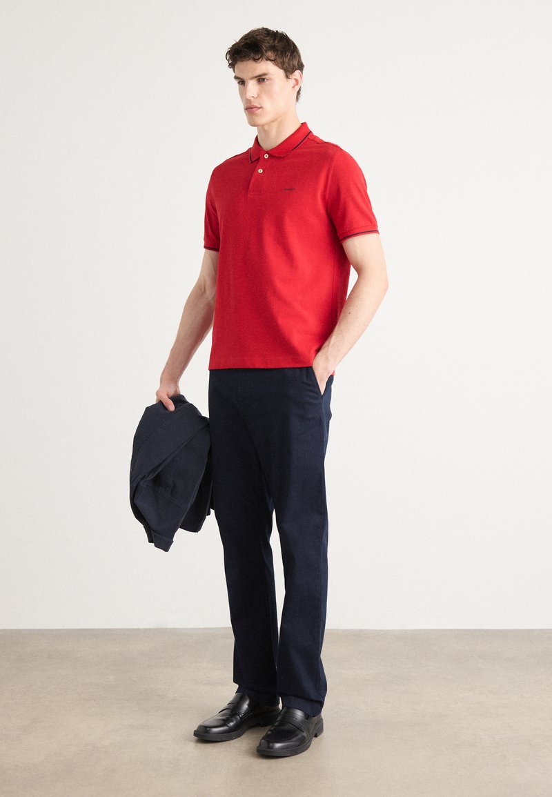 Polo rouge avec un col, des manches courtes et des bordures bleu marine, associé à un pantalon bleu marine et des chaussures noires, tenant une veste bleu marine.